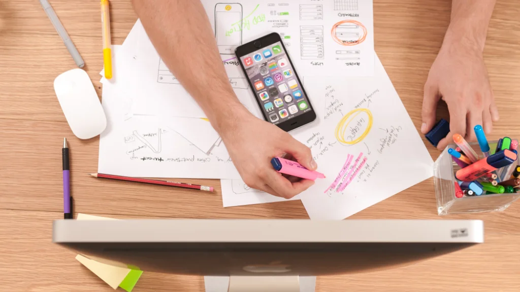Hero Image: Hands on a desk with cell phone, iMac, and school papers.